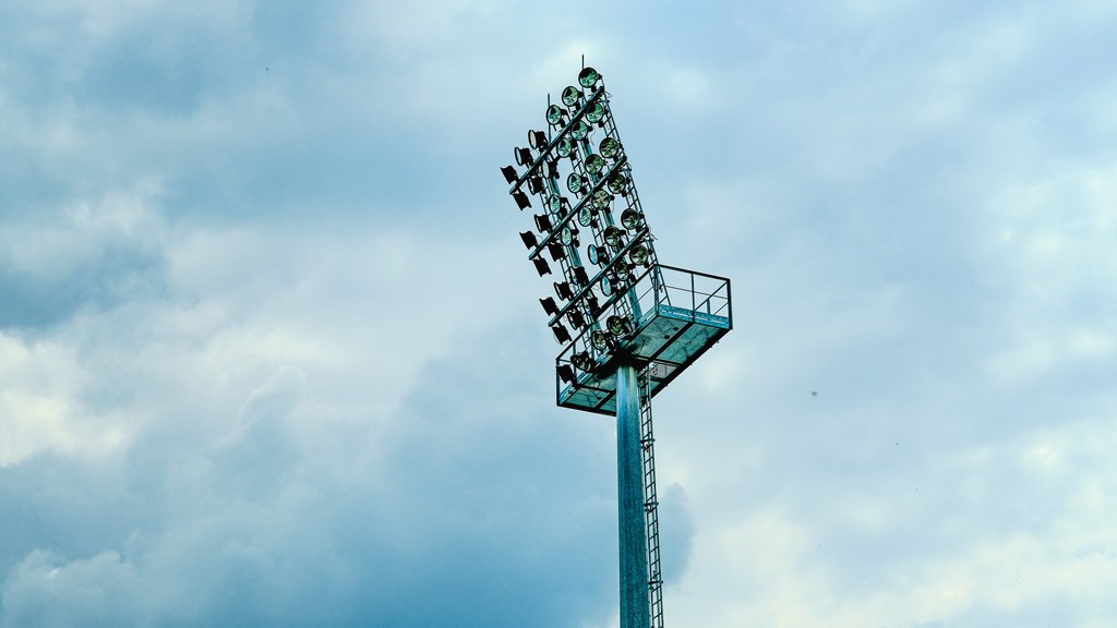 Close-up of a stadium light mast under a cloudy blue sky, creating a dramatic urban scene.