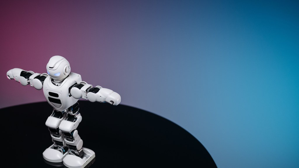 A white humanoid toy robot standing on a reflective black surface in a studio setting with a blue and pink gradient background.