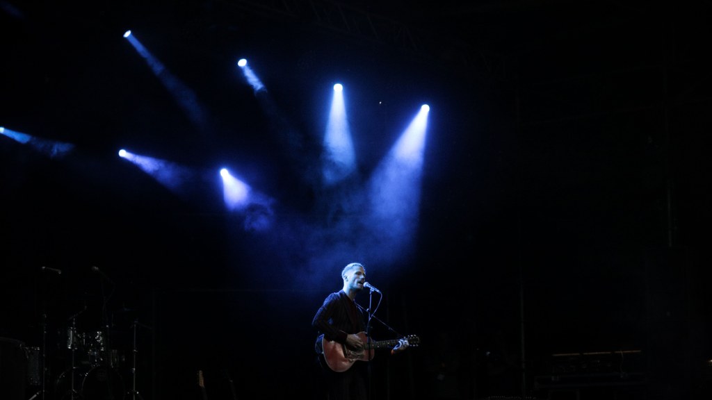 Solo musician performs on stage with guitar under dramatic blue lighting during a live concert.