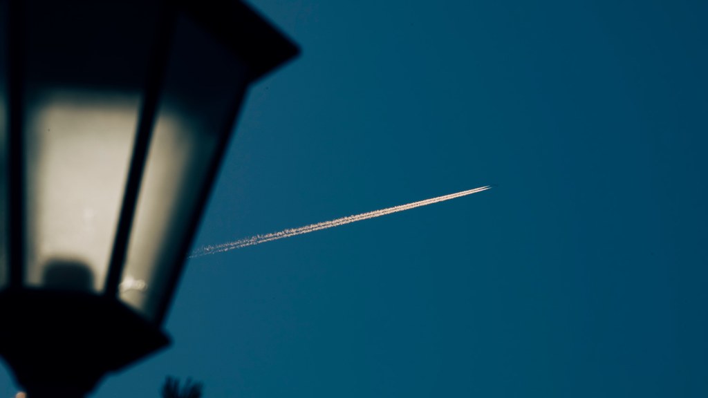 Silhouette of street lamp against the backdrop of a clear sky with an airplane contrail visible.
