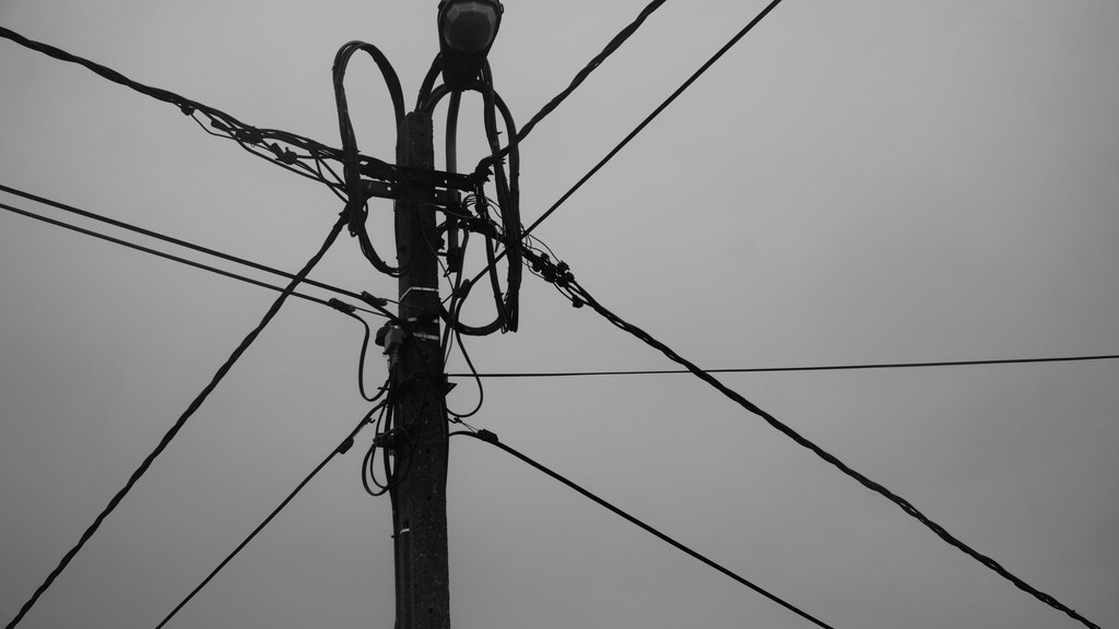 Black and white photo of a utility pole with intersecting power lines and wires.