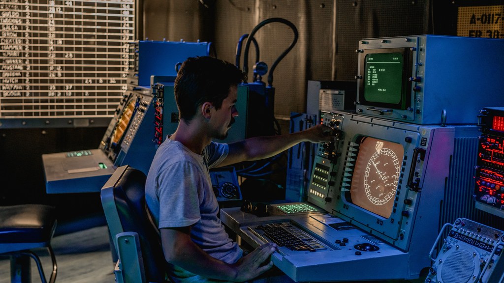 A man working at control panels in a dimly lit industrial room with various monitors.