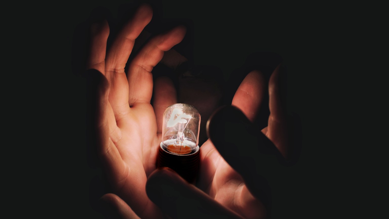 Closeup of anonymous person demonstrating burning lightbulb in palms in middle of darkness
