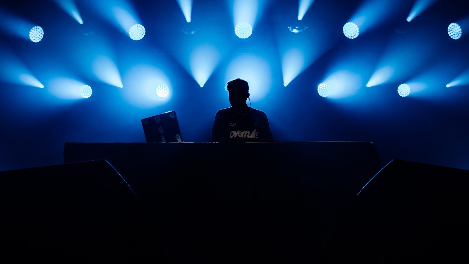 A DJ performs at a music concert, silhouetted against vibrant blue stage lights.