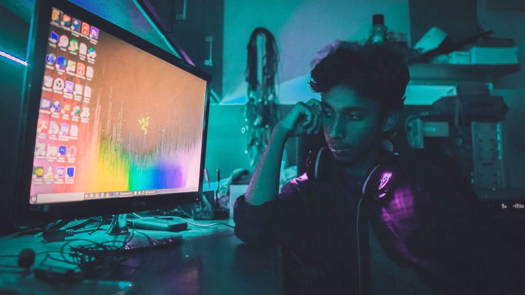 A young man wearing headphones sits by a computer in a dimly lit room, deep in thought.