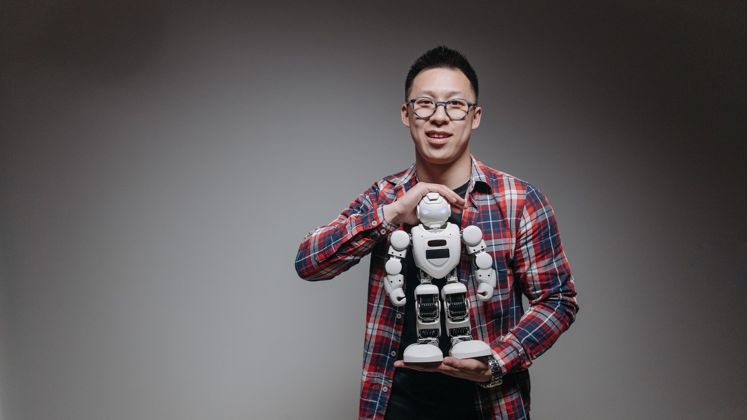 Asian man with eyeglasses holding a toy robot in a studio with a gray background.