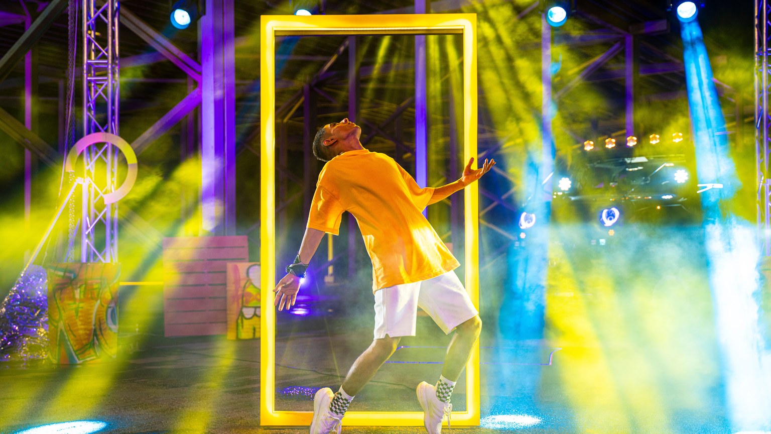 A man dances energetically on stage with colorful lights and smoke in a vibrant setting.