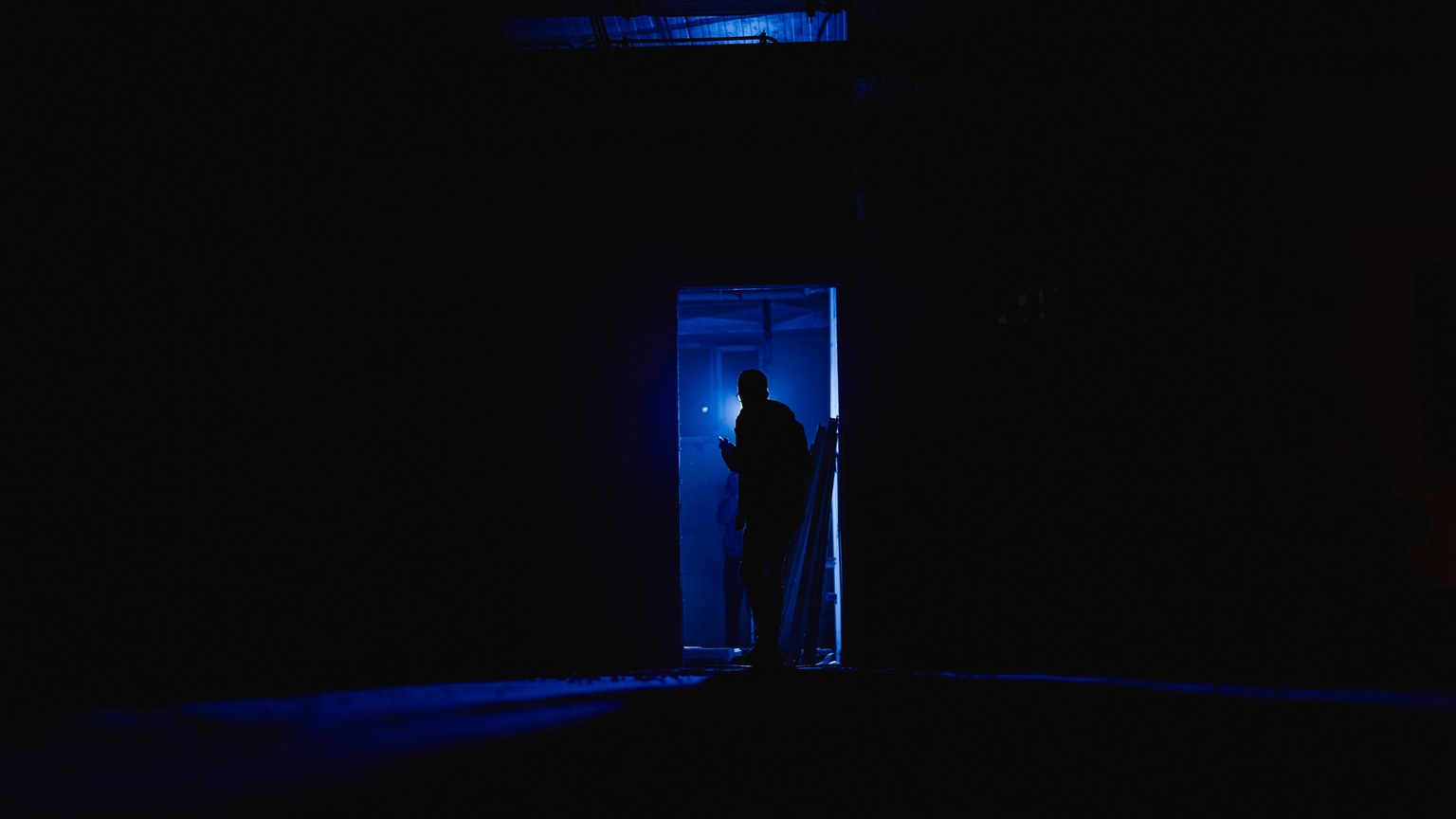 A mysterious silhouette stands in a dimly lit room, bathed in blue light, suggesting intrigue and solitude.