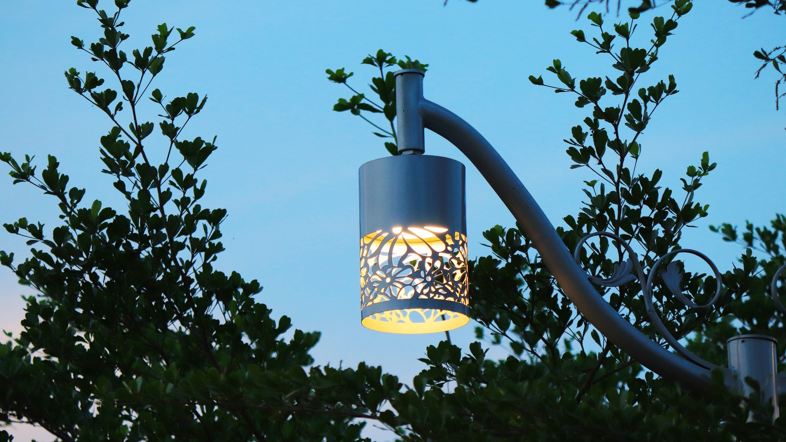 Elegant street lamp glowing against a backdrop of lush green leaves at dusk.