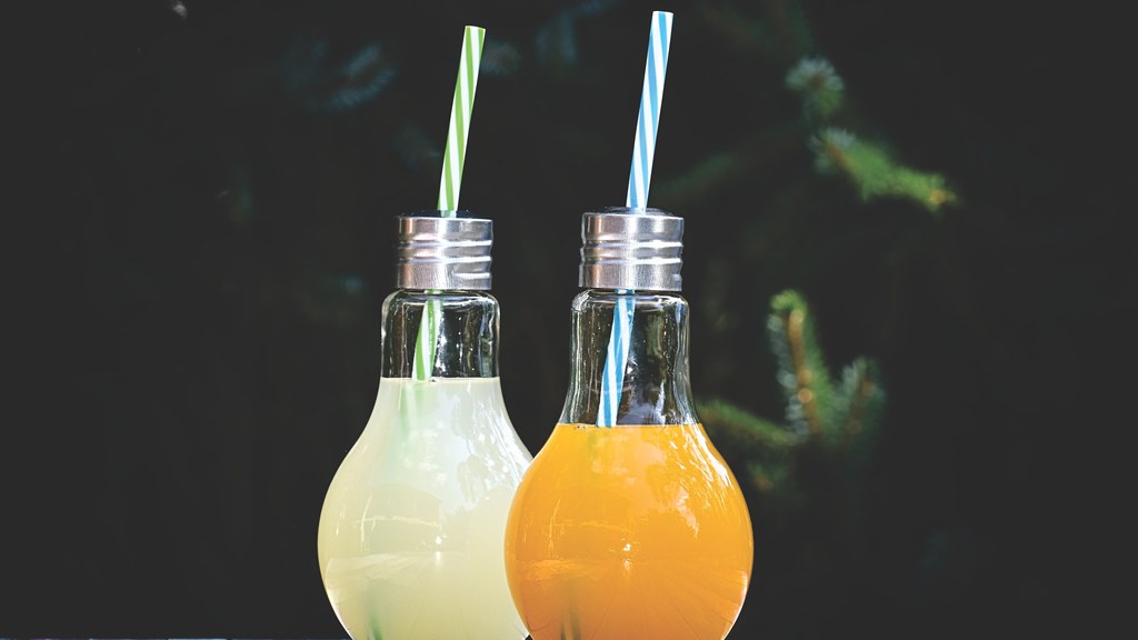 Two vibrant fruit juices in lightbulb-shaped glasses with straws on a colorful table.