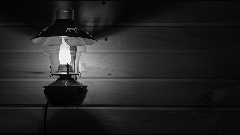 Black and white photo of a vintage lamp illuminated against a wooden wall interior setting.