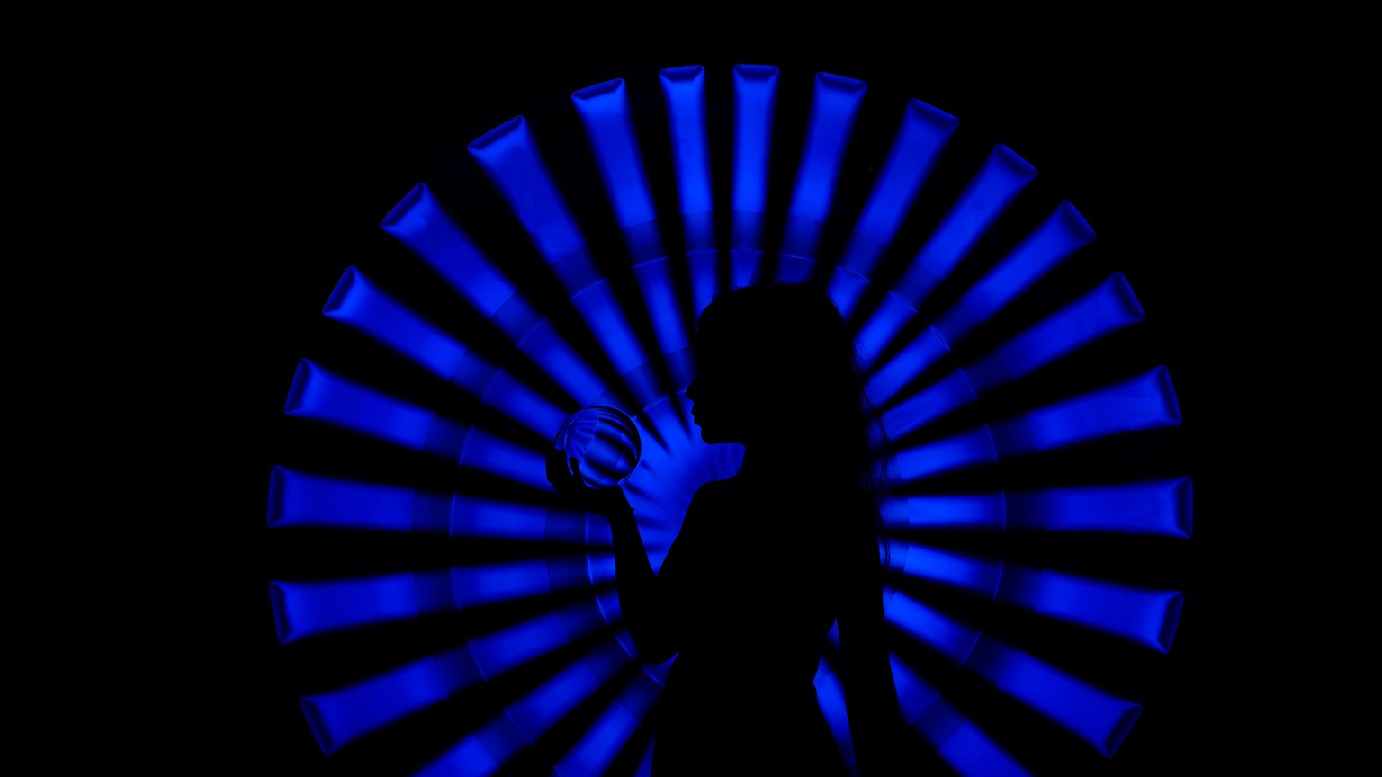 Side view silhouette of unrecognizable female with long hair standing in dark room with crystal ball in hand near bright neon blue illumination