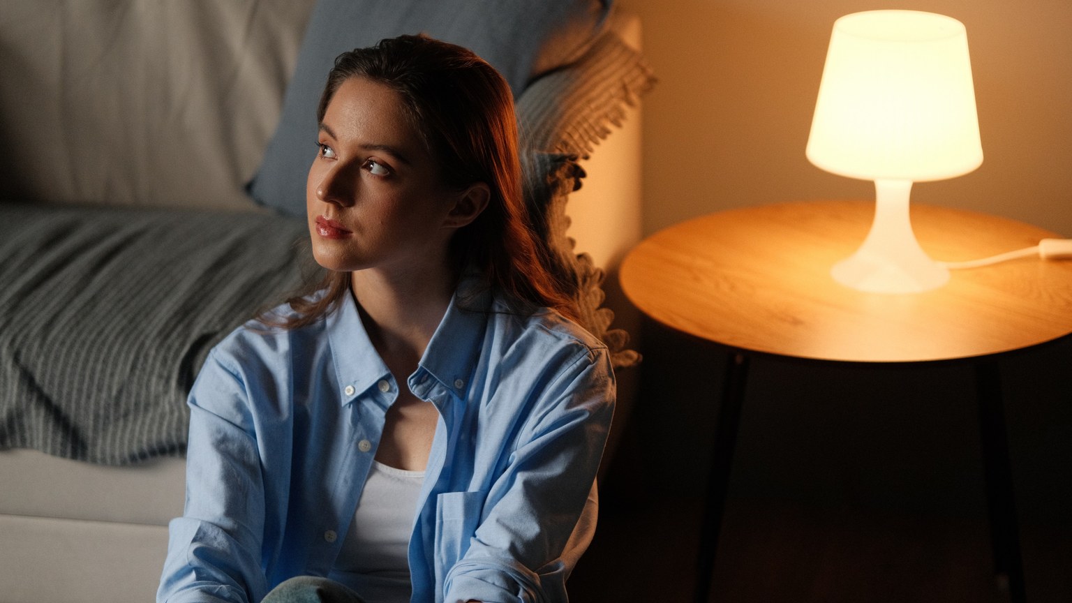 Young woman in casual attire sits thoughtfully by a warm-lit lamp indoors.