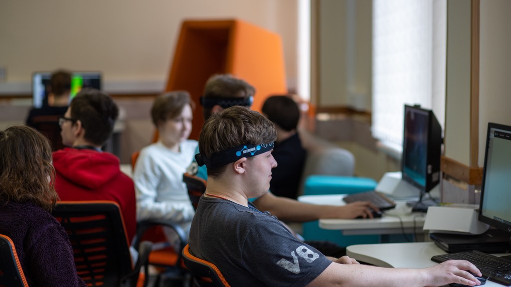 Group of teenagers engaged with computers and headsets in a modern classroom.