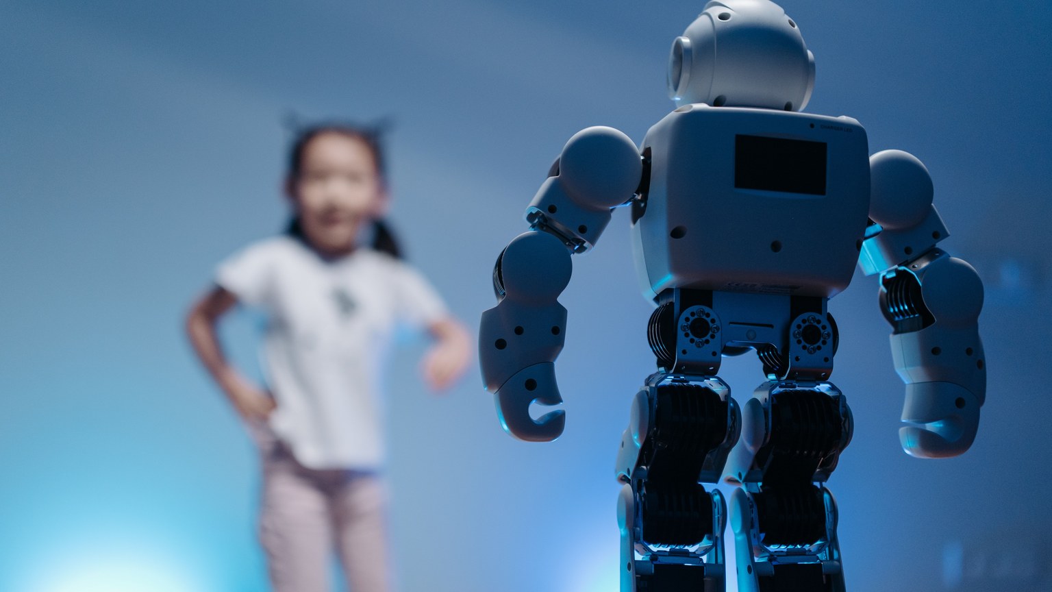 A young girl playfully interacts with a humanoid robot in a futuristic indoor environment featuring soft blue lighting.