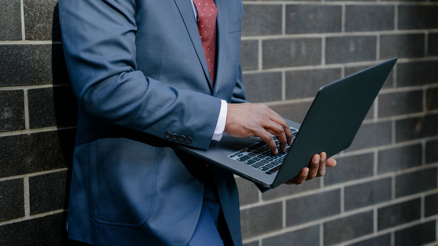 Mid-adult businessman in blue suit typing on laptop against brick wall outdoors.