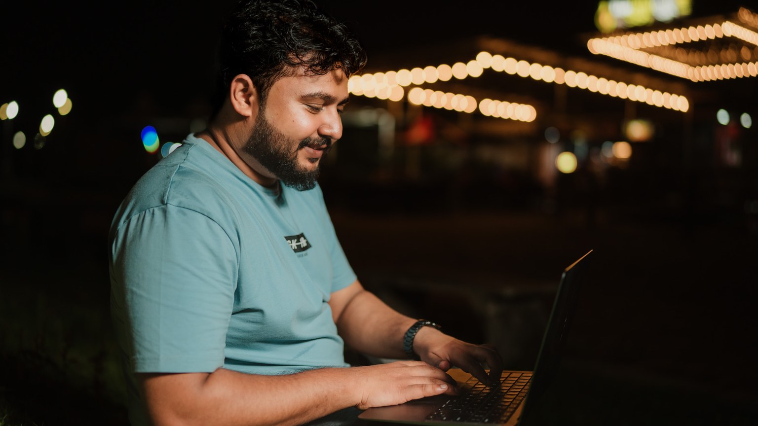 A man in a blue t-shirt typing on a laptop outdoors under string lights at night.