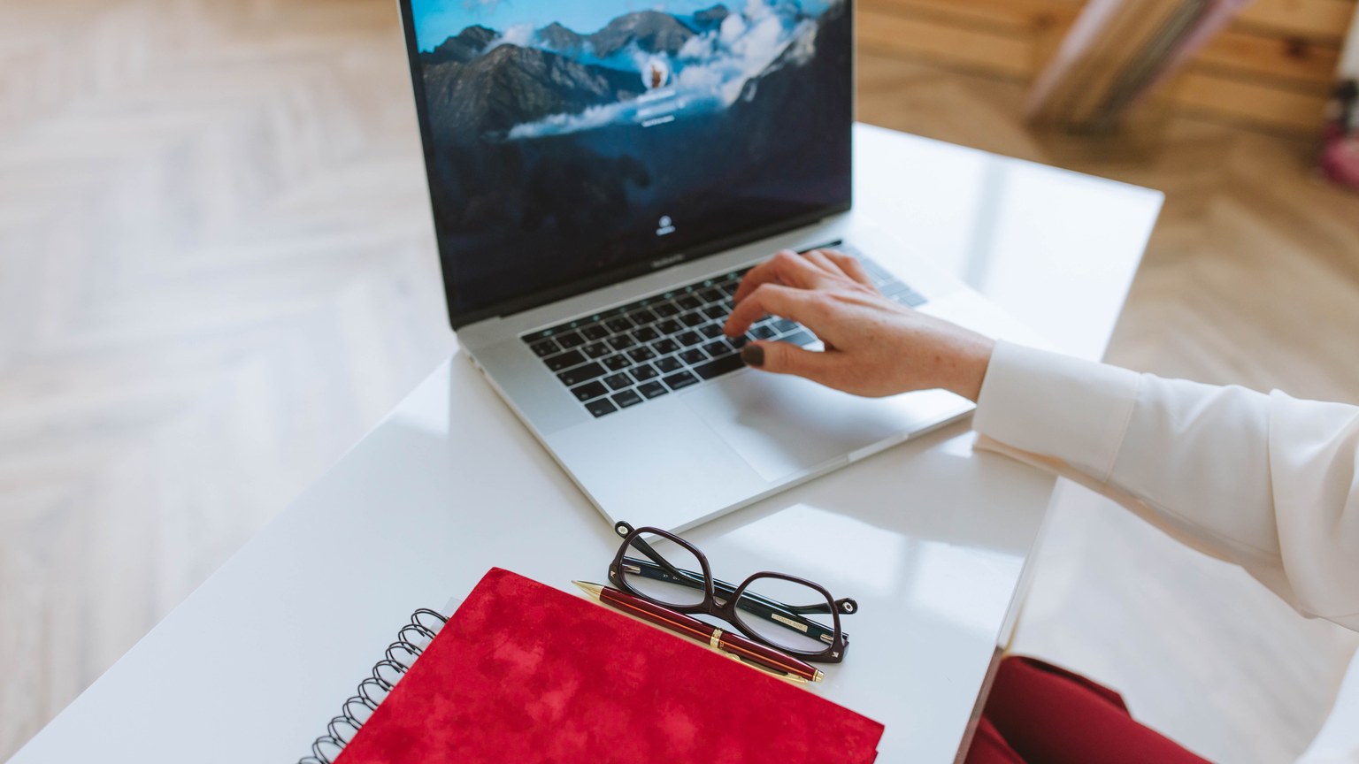 A person working on a laptop with a red notebook and glasses on a white table.