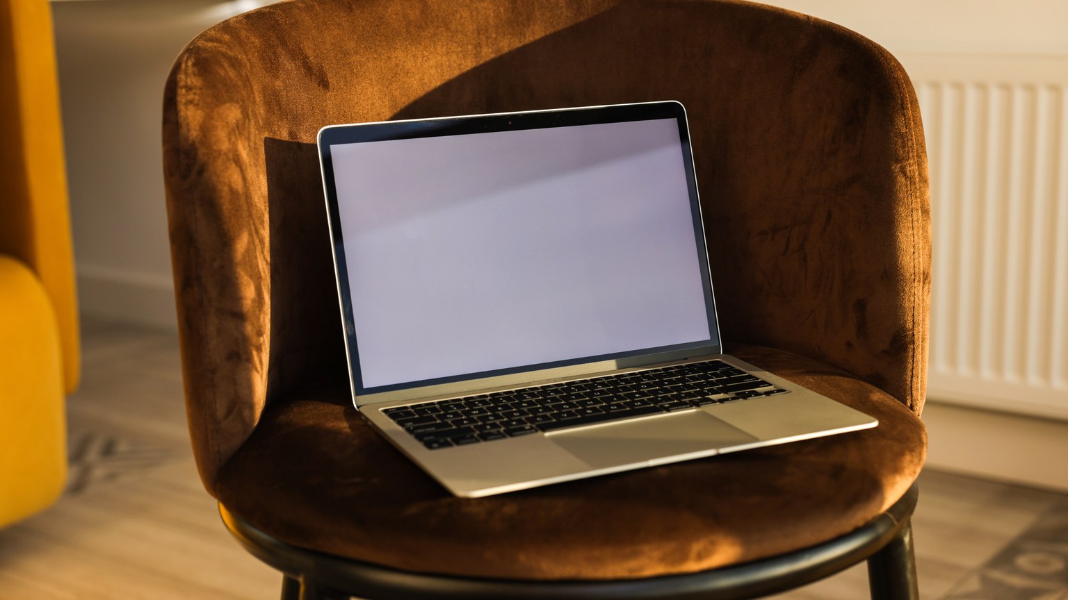 Laptop with a blank screen placed on a brown velvet chair in a bright room.