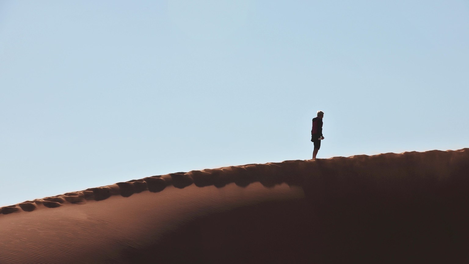 A lone hiker walking on a vast dune at Sossusvlei, Namibia, against a clear sky.