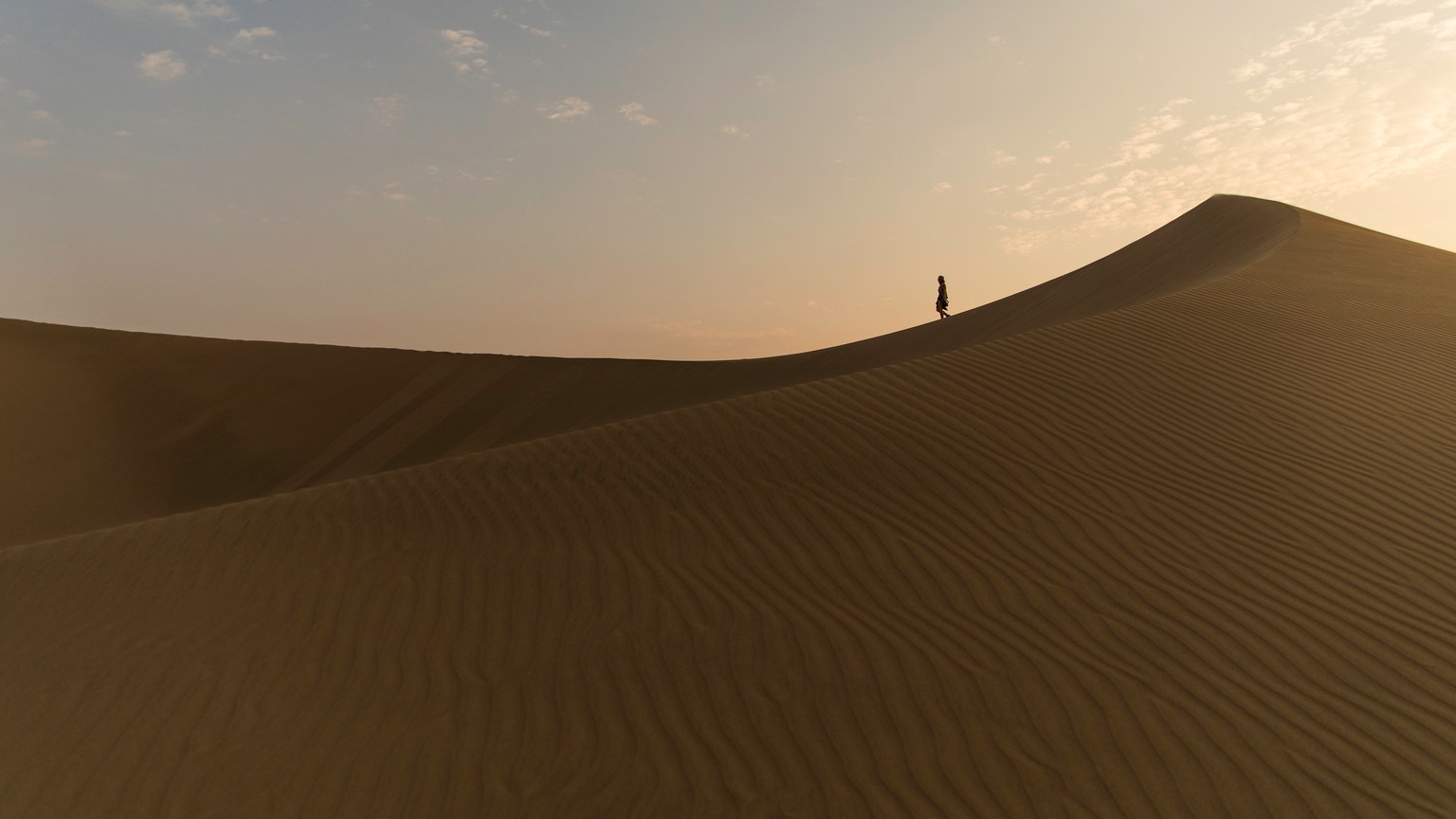 Silhouette of a person walking on a vast sand dune in the desert at sunset in Huacachina, Peru.