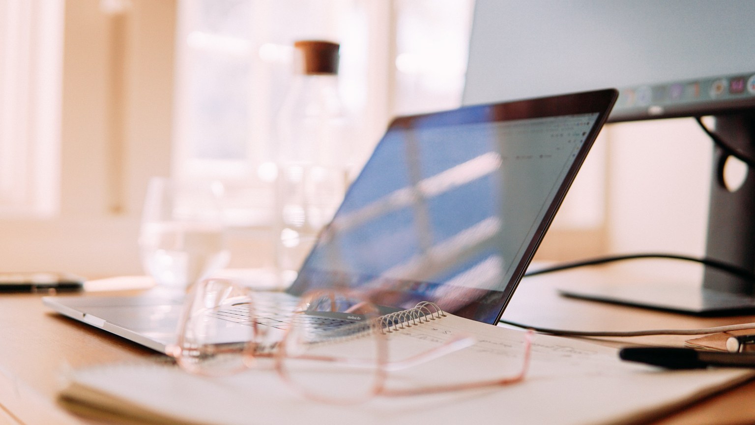 A well-lit home office setup featuring a laptop, monitor, and glasses, ideal for remote work.