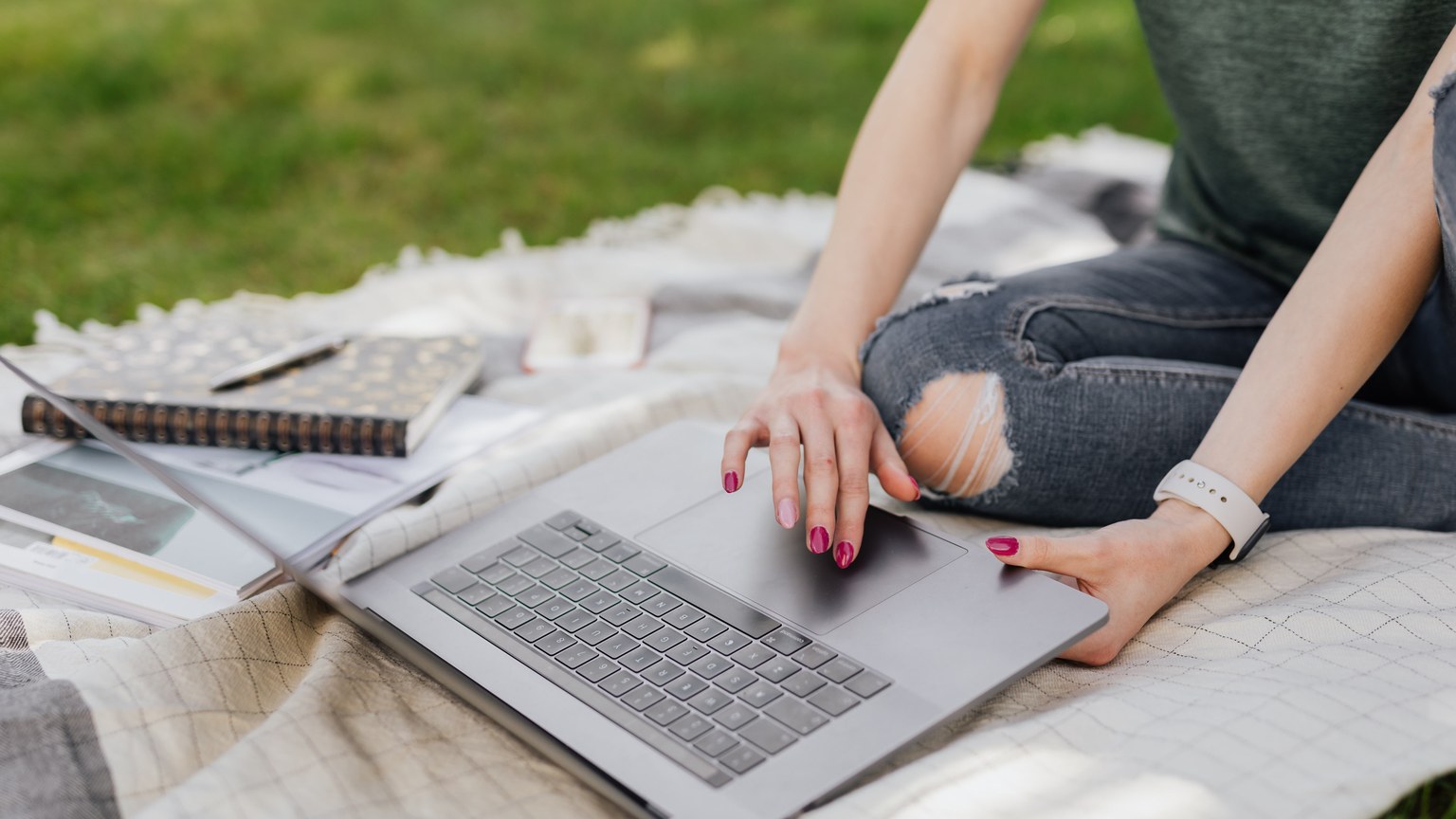 Crop unrecognizable female student doing Internet research on laptop while sitting on cozy blanket in green summer park on sunny day