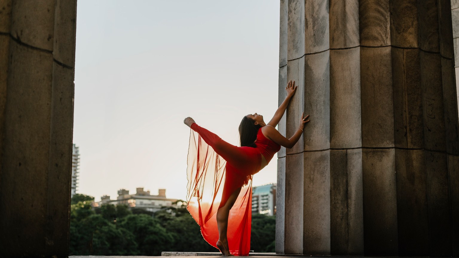 A graceful female dancer in a red dress striking a pose between city pillars during dusk.