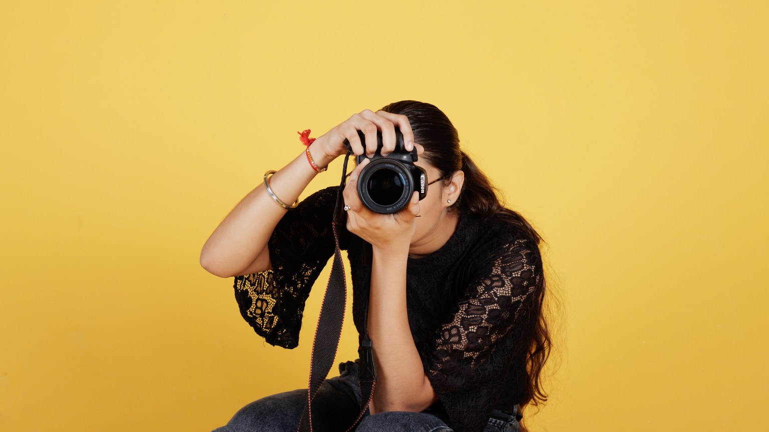 A young female photographer taking a photo with a DSLR camera against a yellow background.