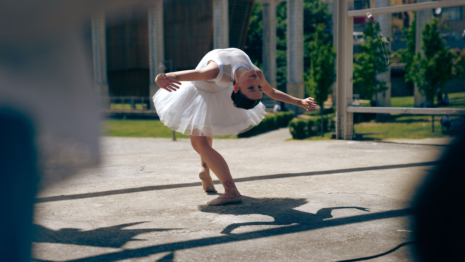 Graceful dancer in white tutu performs ballet outdoors, casting a shadow on pavement.