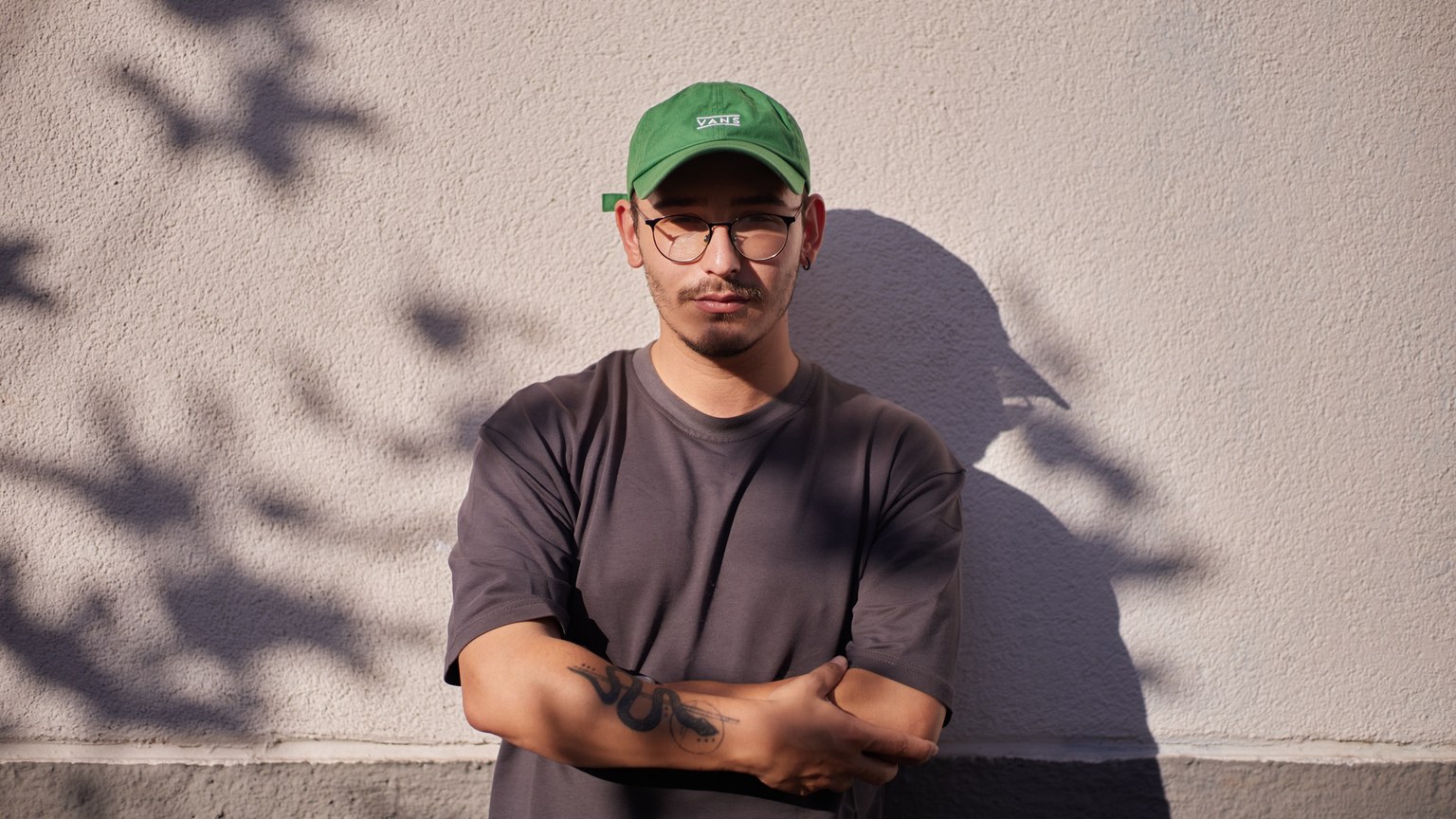 Man wearing green cap and glasses stands against a wall, casting shadows in the sunlight.