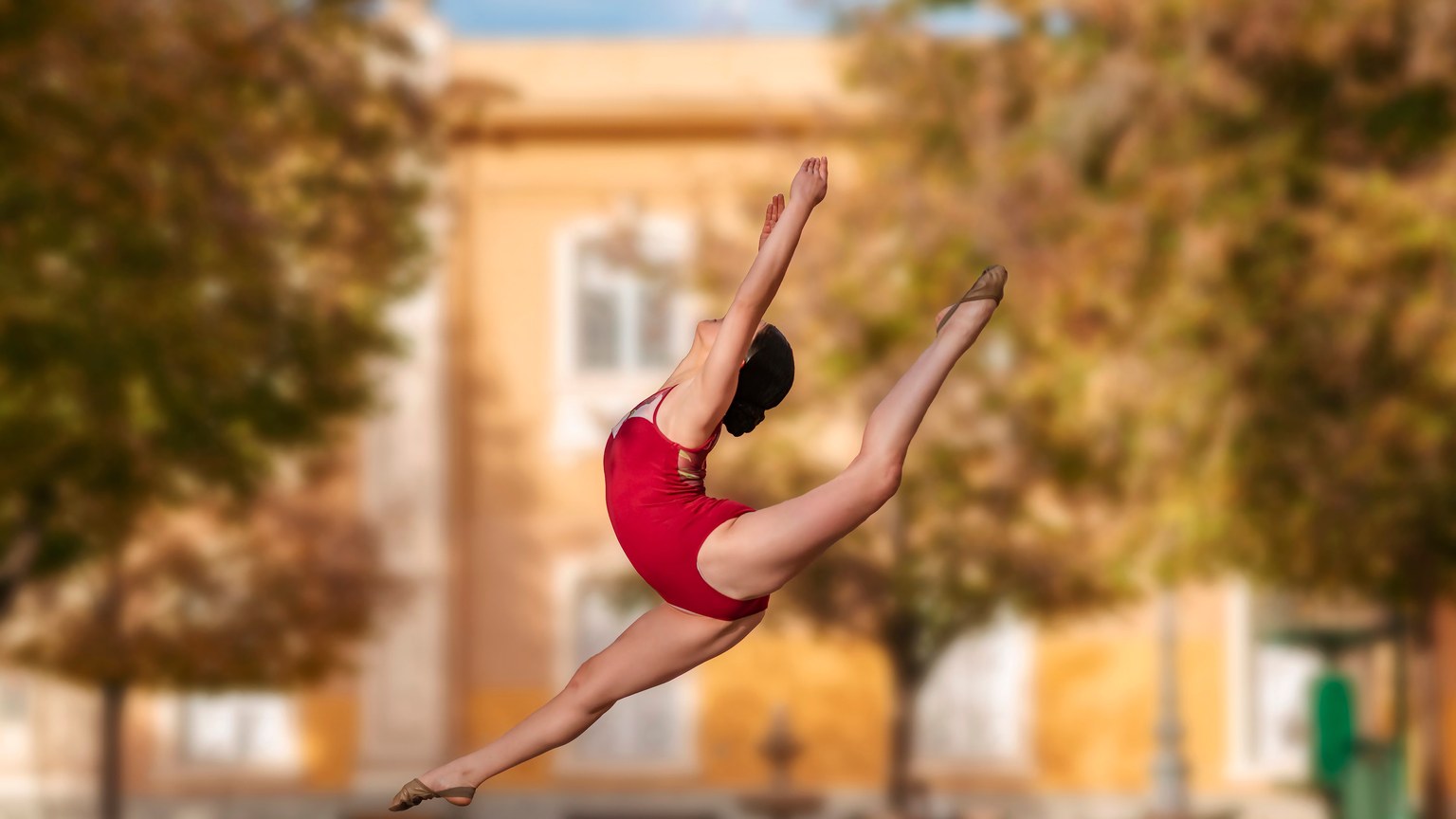 Aerial view of a female dancer in a red leotard performing a mid-air split outdoors.