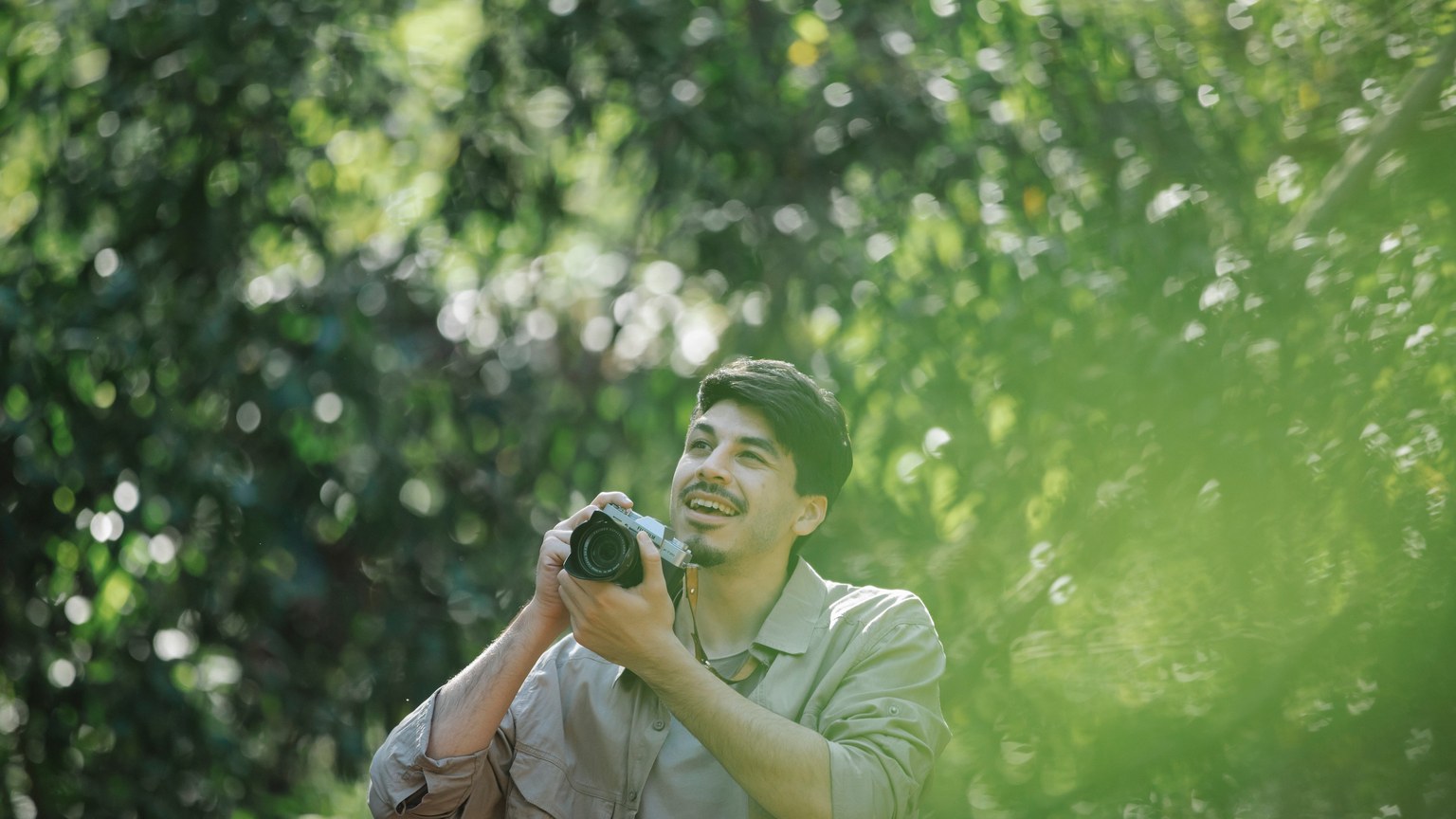 Happy young bearded male photographer in casual clothes with retro photo camera looking up against green trees in park