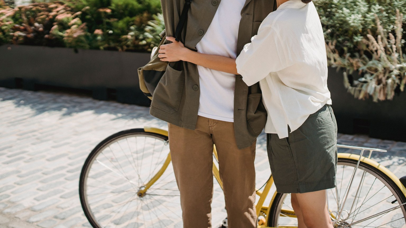 Unrecognizable loving couple in casual wear embracing while standing on pavement near bicycle and green plant in city on sunny street