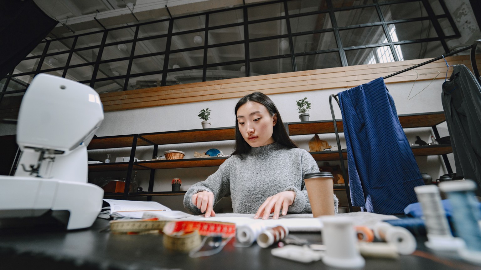 A focused fashion designer works in a studio, surrounded by sewing tools, cloth, and a coffee cup.