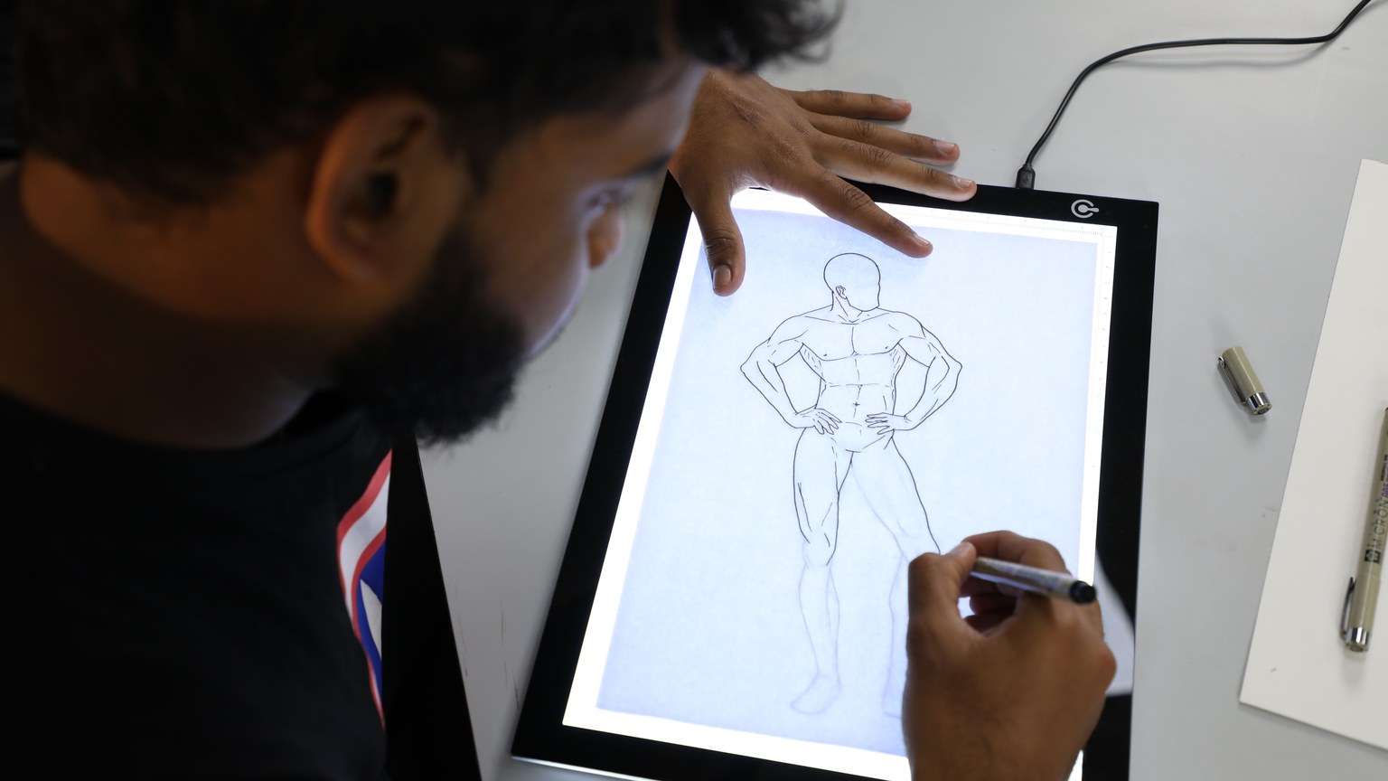 Illustrator sketching a superhero figure on a digital lightboard in an art studio setting.