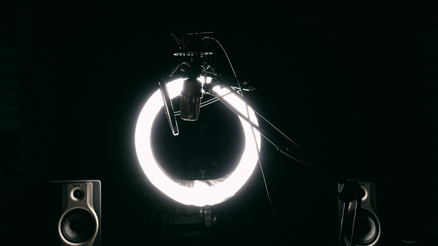 Dark studio room with condenser microphone, ring light, and speakers for recording purposes.