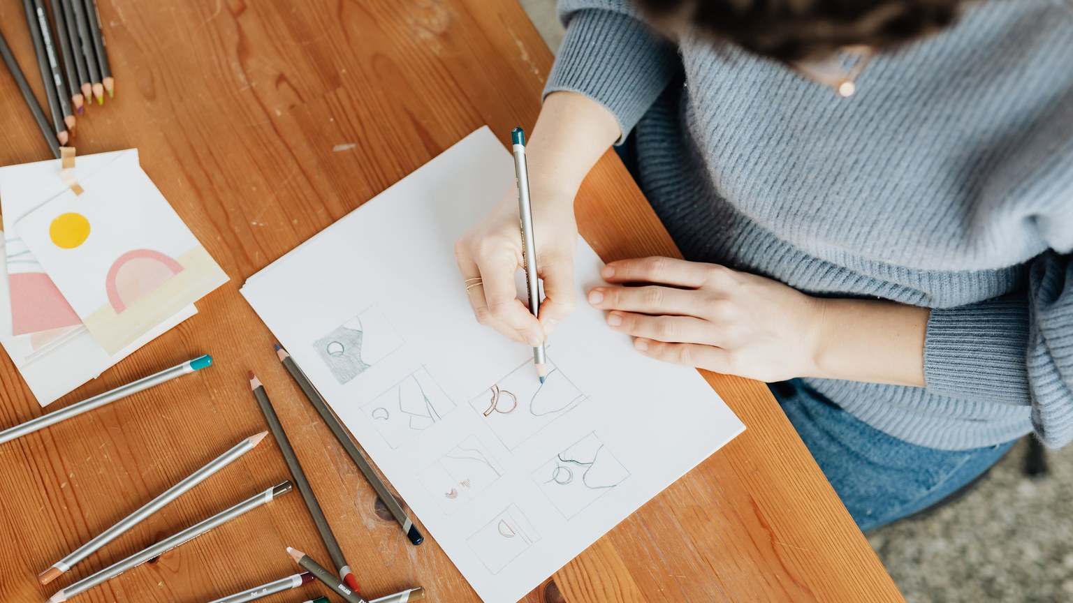 Top view of crop anonymous female illustrator in sweater and jeans with eyeglasses making sketch with pencil on paper while sitting at wooden desk at work