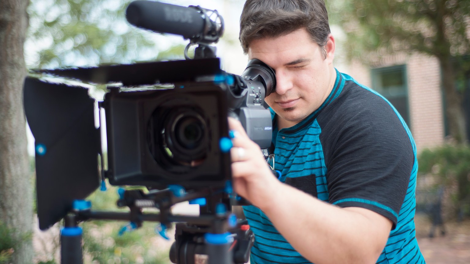 Young male videographer using a professional video camera outdoors with a blurred background.