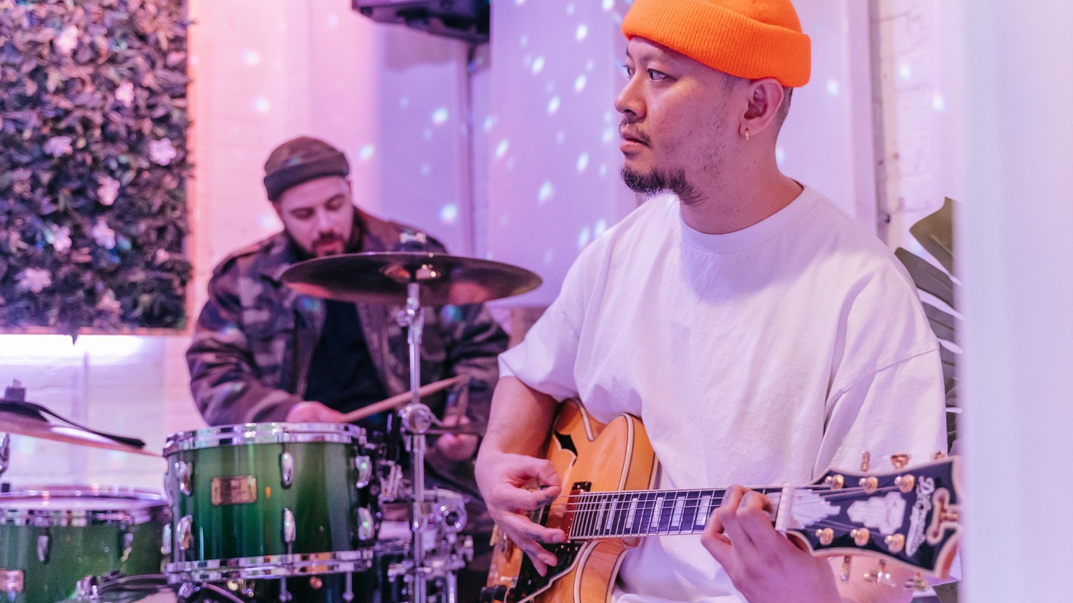 Two musicians playing guitar and drums indoors in a vibrant setting with orange beanie and green drum set.
