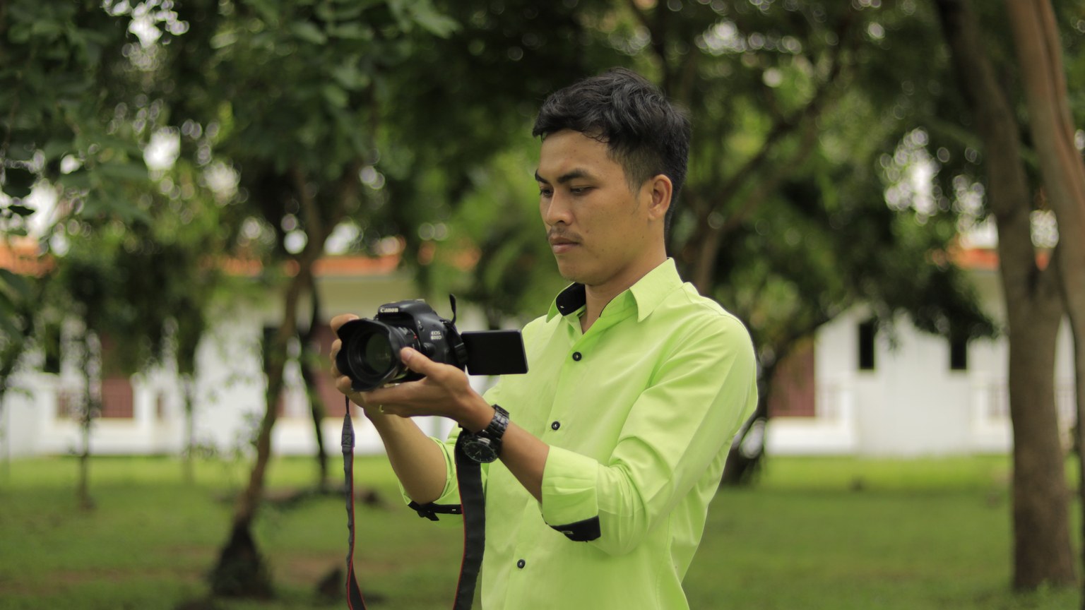 Asian male photographer in green shirt using camera outdoors in lush setting.