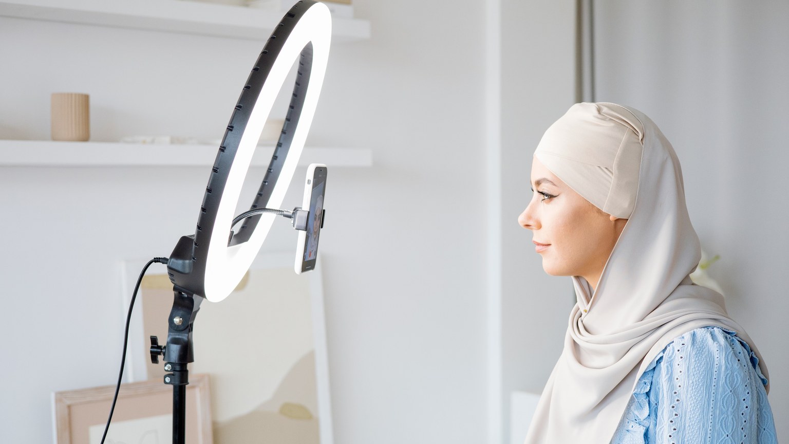 A woman in hijab uses ring light for video content creation indoors.