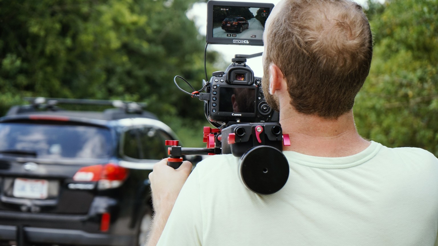 Videographer using shoulder-mounted camera rig to film vehicle scene outdoors in daylight.
