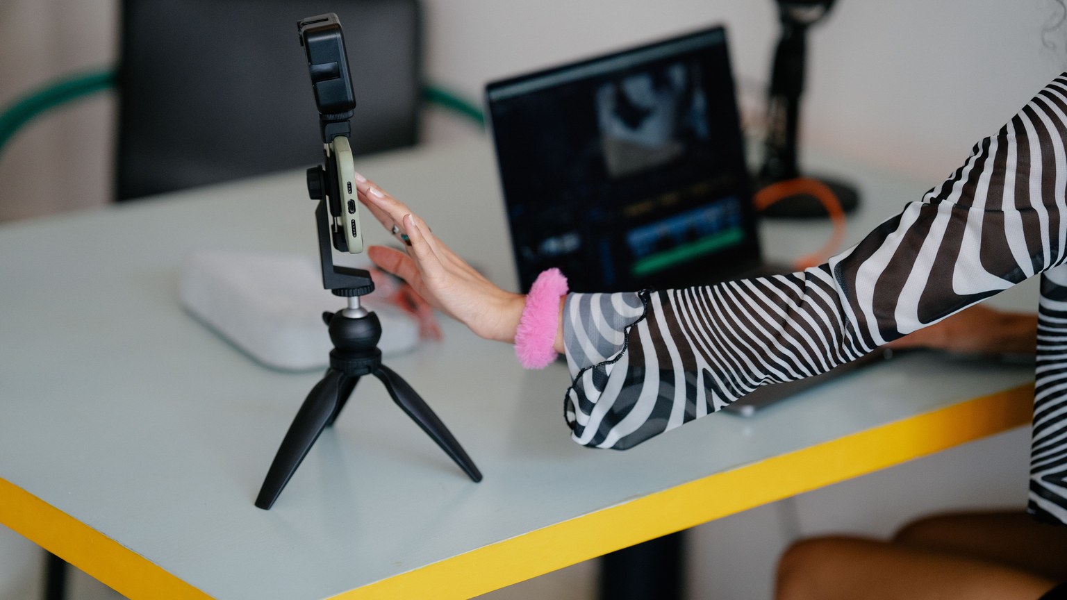 Woman adjusting smartphone on tripod with laptop setup for video editing.