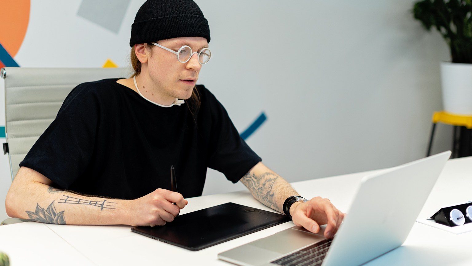 Tattooed designer working on a laptop and tablet in a stylish office setting.