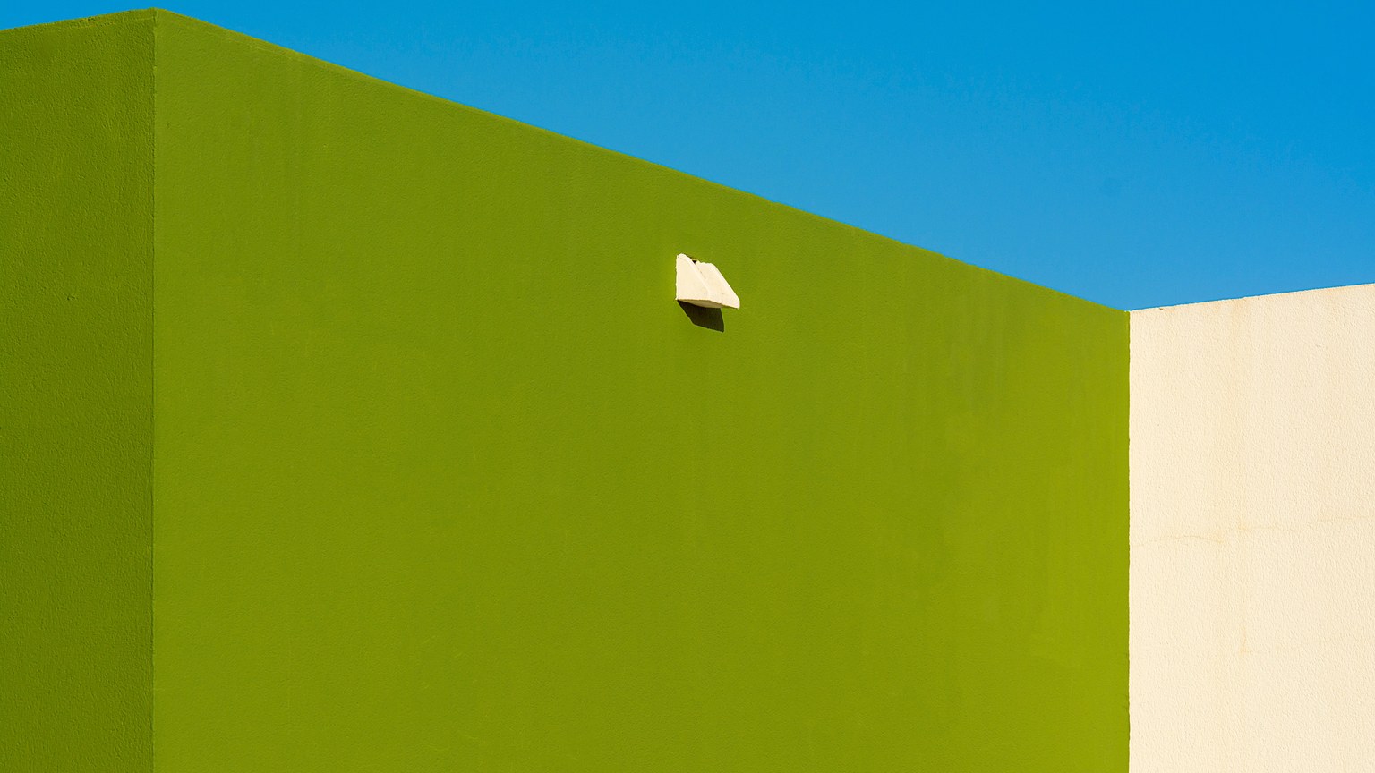 A minimalist composition featuring a green wall with a protruding fixture against a deep blue sky.