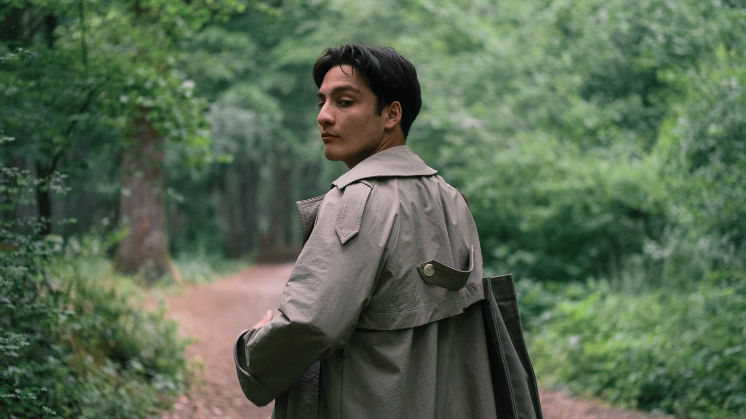A contemplative scene of a man in a trench coat walking through a lush green forest.