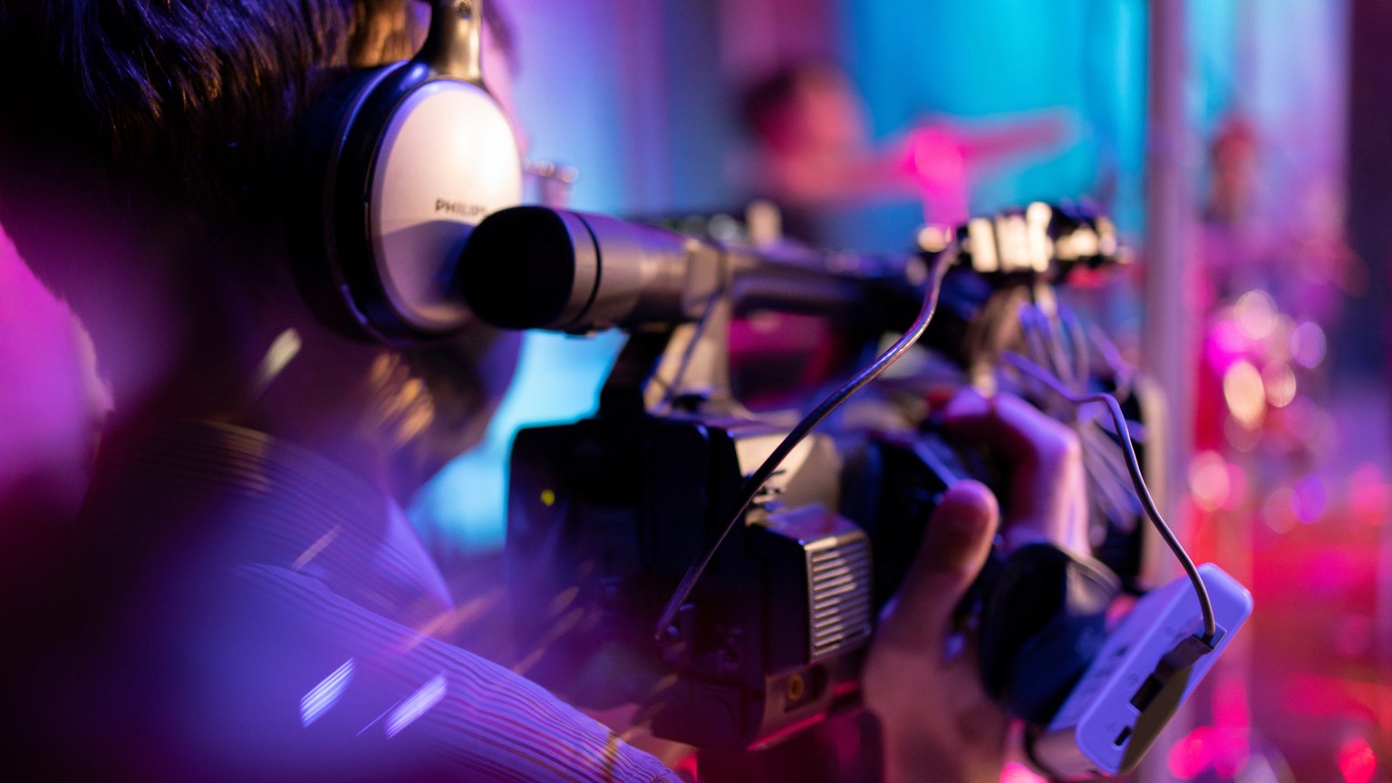 A videographer with headphones focuses intently while filming an indoor event under colorful lights.