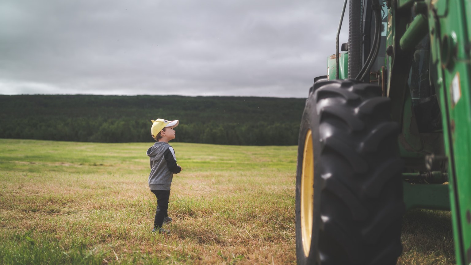 A young child observes a large green tractor in a vast open field under an overcast sky.