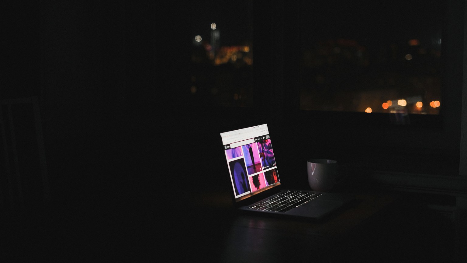A laptop glows in a dark room at night with a cityscape through the window. Ideal for tech and solitude themes.